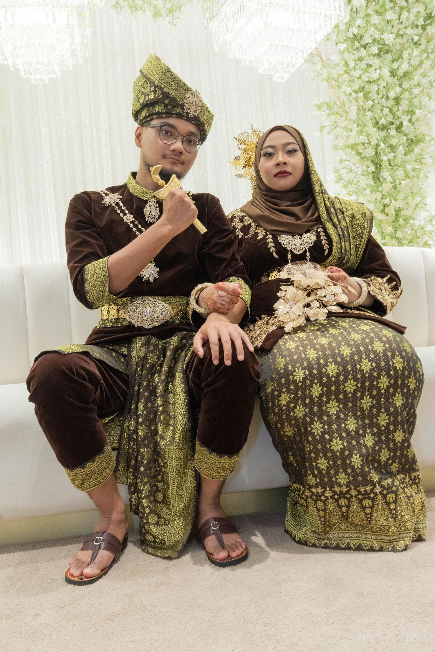 Couple in ornate brown and gold traditional Malay costumes seated on white sofa.