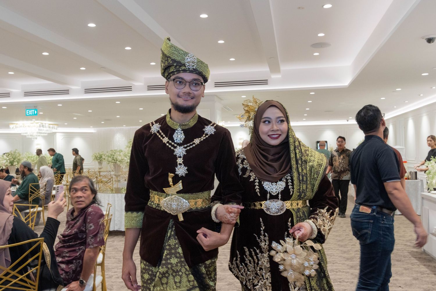 Couple in matching dark traditional Malay formal wear at an indoor celebration.