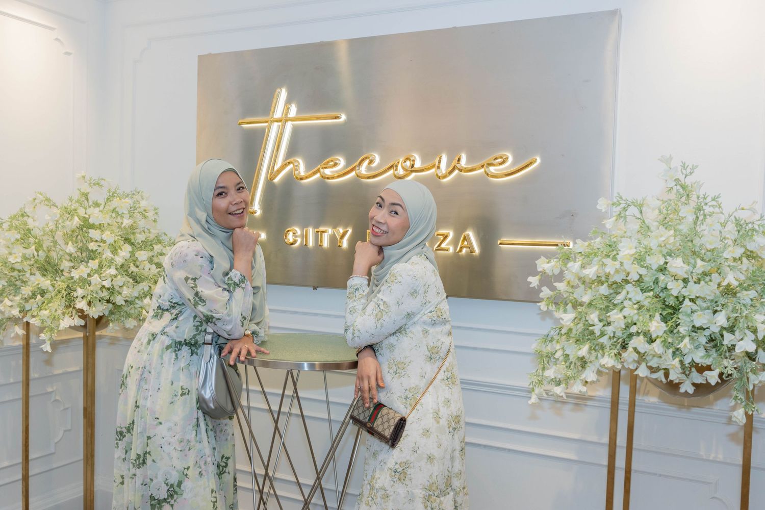 Two people in floral printed dresses pose in front of an illuminated 'Treasure' sign with white flower decorations.