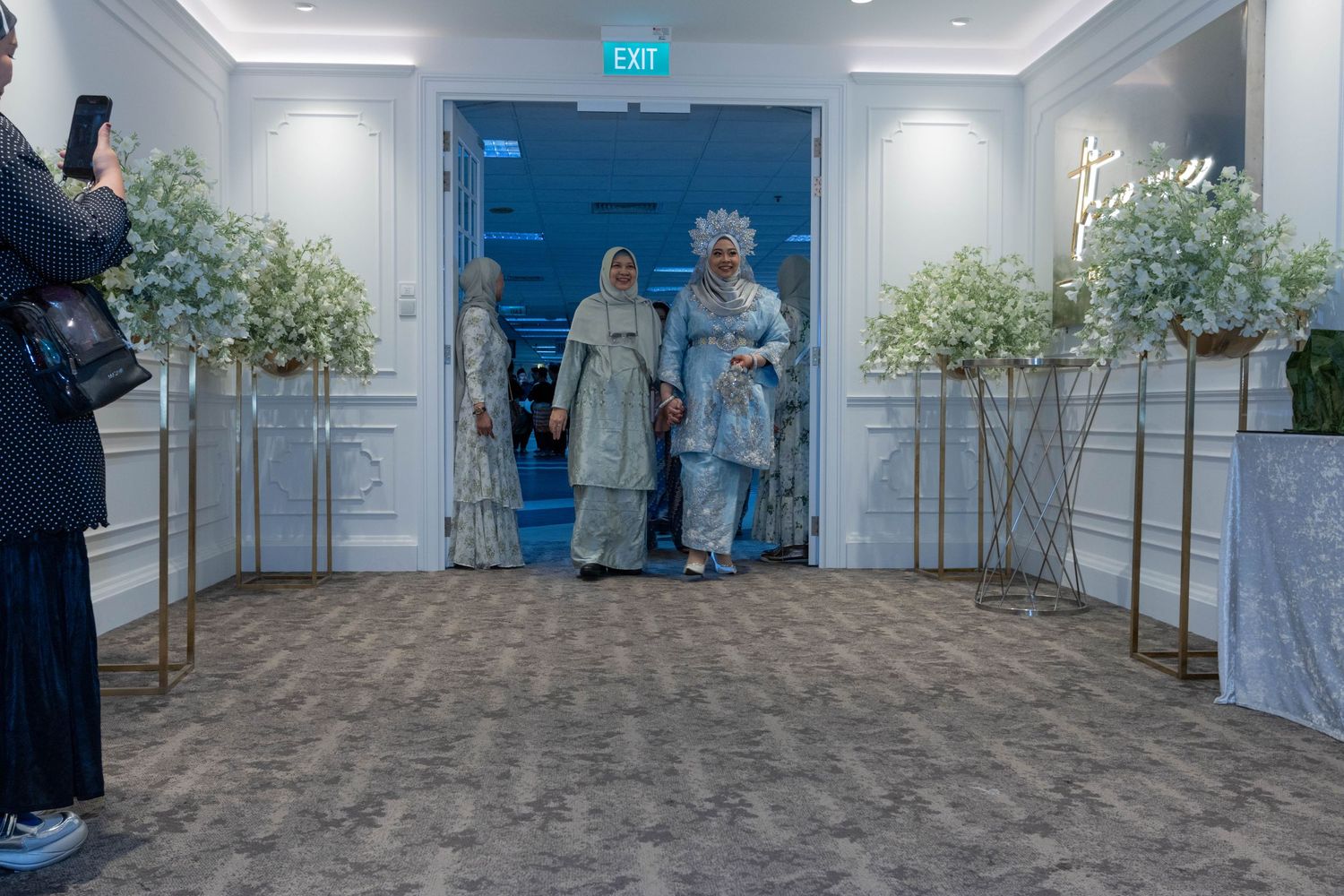 People in formal attire enter through blue doors flanked by white floral arrangements in an elegant venue.