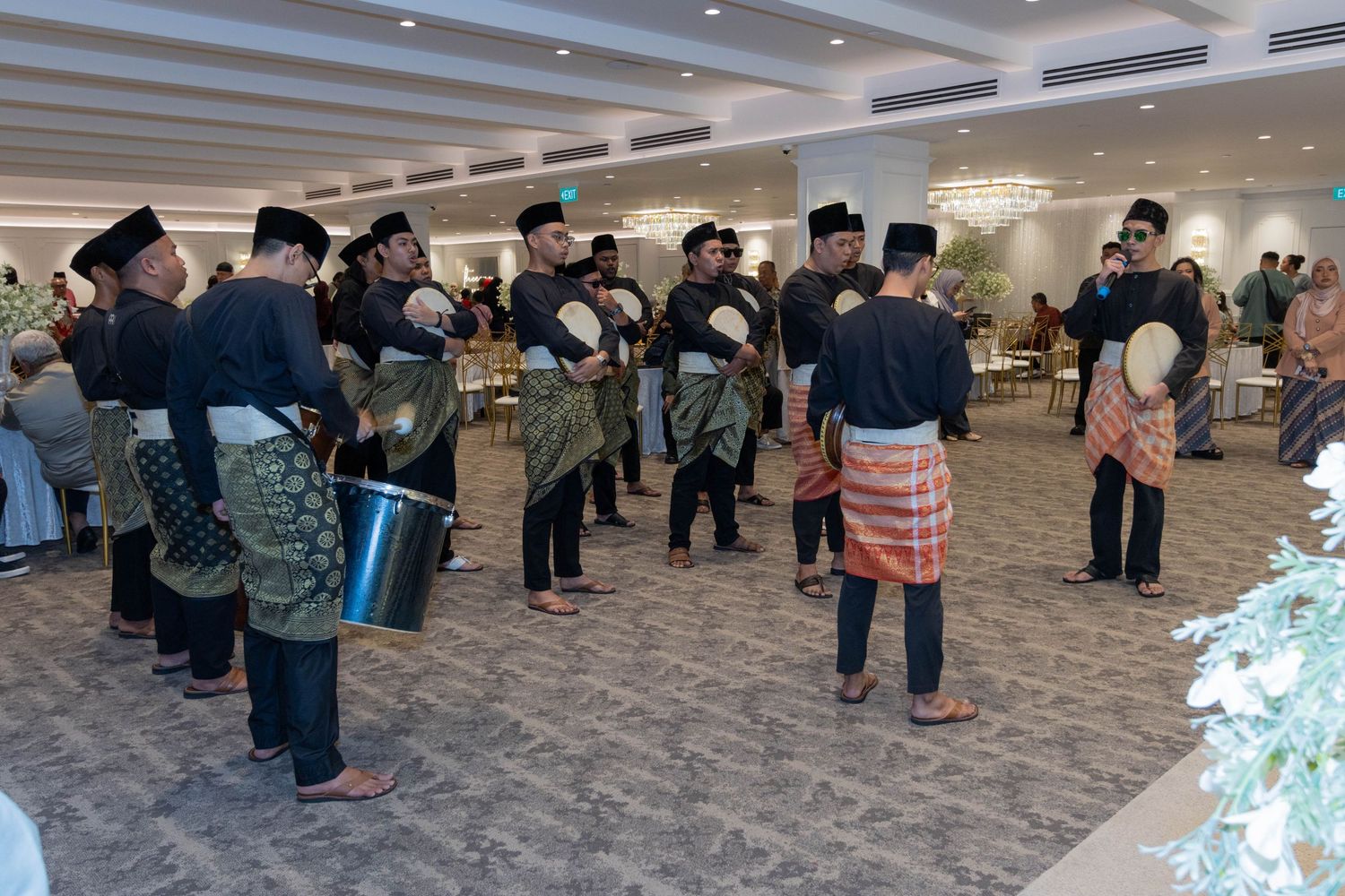 Group of traditionally dressed musicians with drums gather in formal event space.