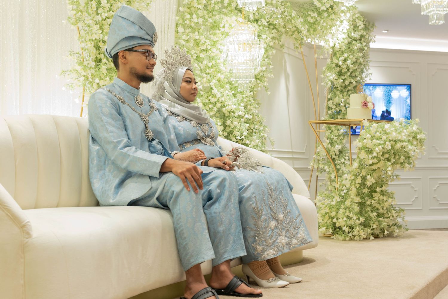 Couple in matching powder blue traditional outfits seated on white couch with floral decorations.