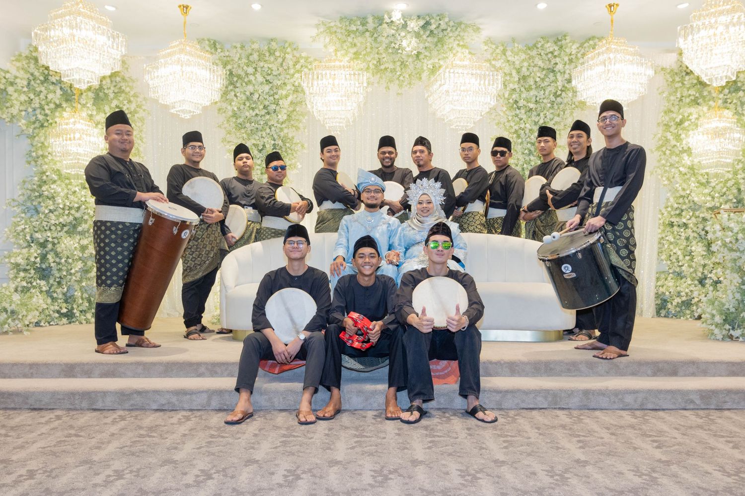 Traditional musicians with drums pose for group photo against floral backdrop.