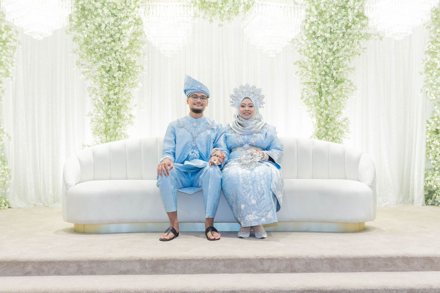 Couple in coordinated light blue attire seated on white couch against floral wall backdrop.