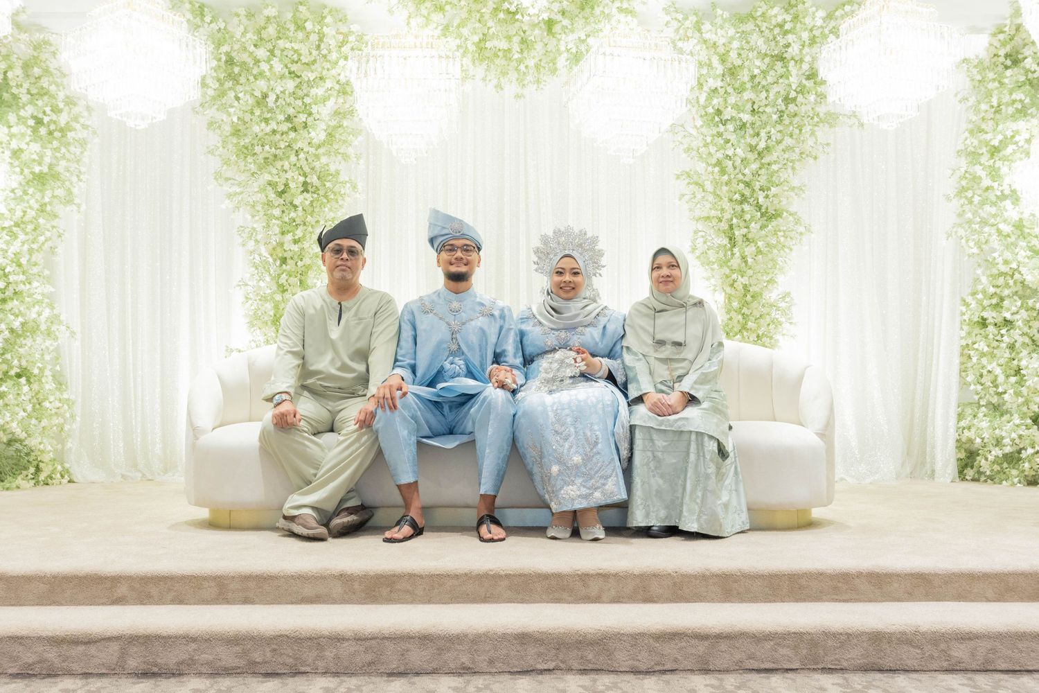 Small group in formal traditional attire seated on white couch against floral backdrop.