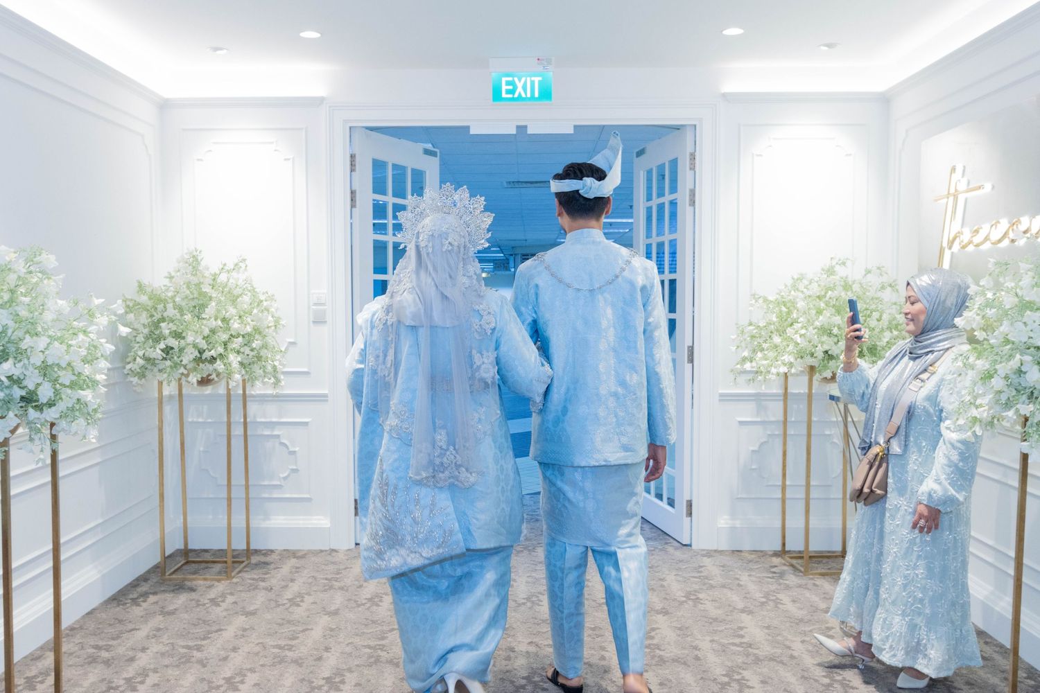 A couple in matching light blue traditional outfits walk down an elegantly decorated white hallway.