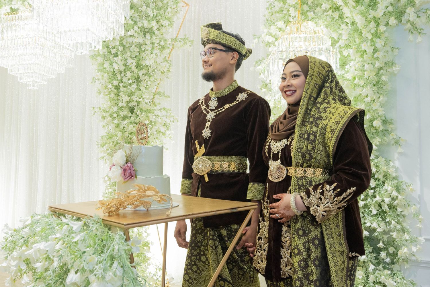 A couple in traditional Malay wedding attire featuring dark velvet and gold embellishments.