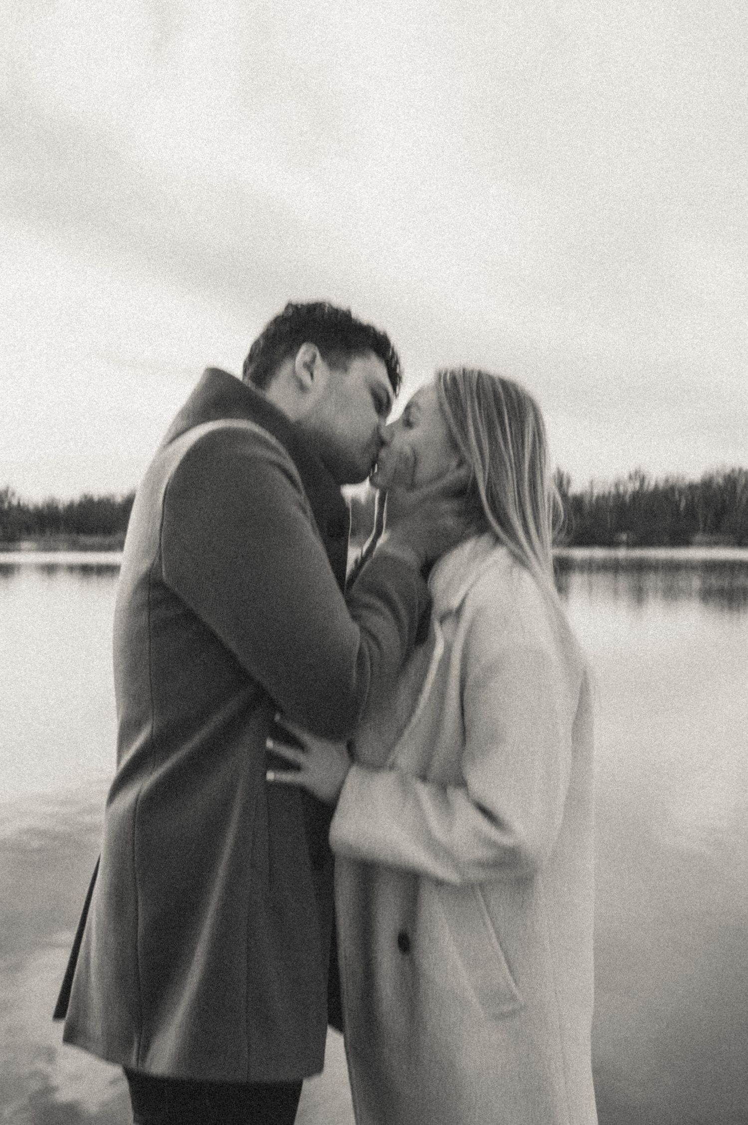 A couple shares tender moments by a serene lake on an overcast day.