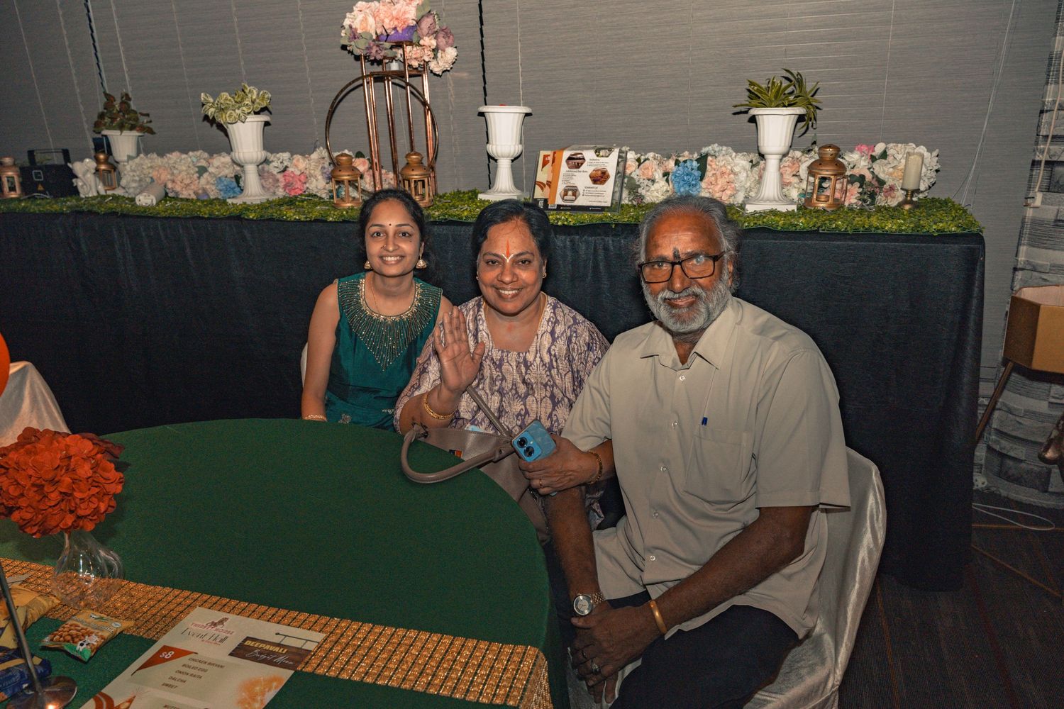 Guests seated at a decorated table with floral arrangements and green tablecloth at an indoor celebration.
