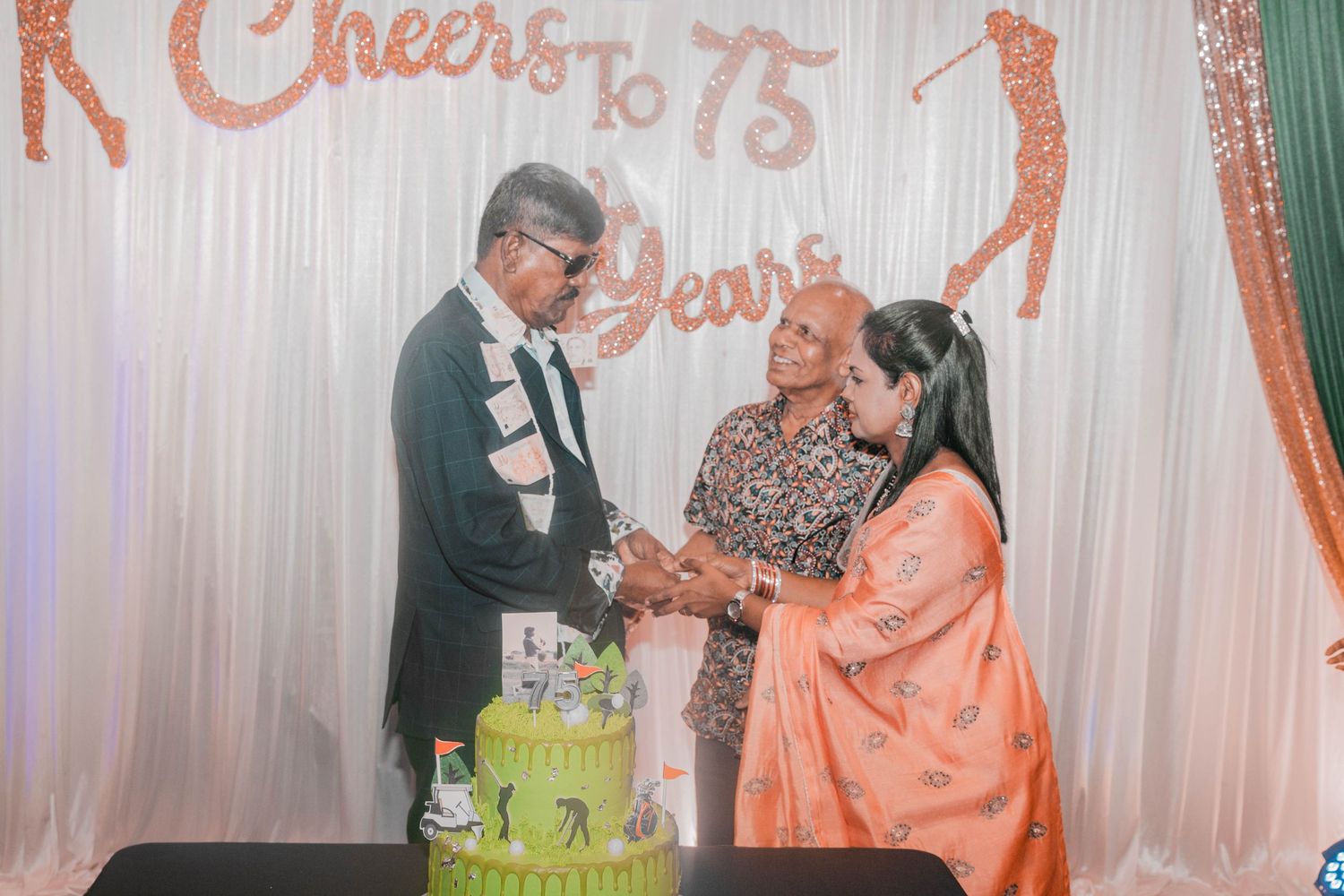 People gathered around a green celebration cake at a 75th anniversary party with decorative backdrop.