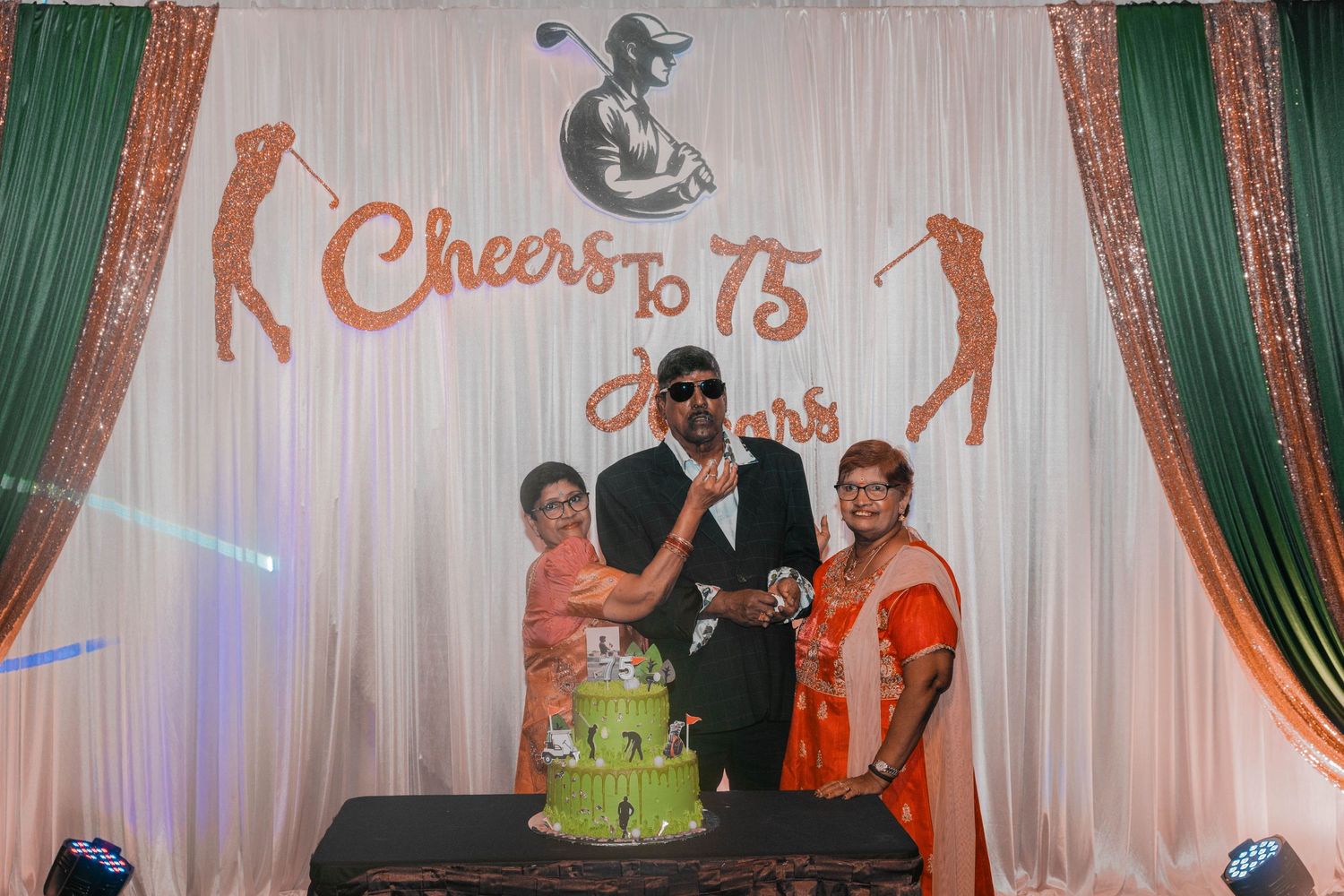 Group celebrates around a green cake at a 75th anniversary party with sparkly backdrop and decorative elements.