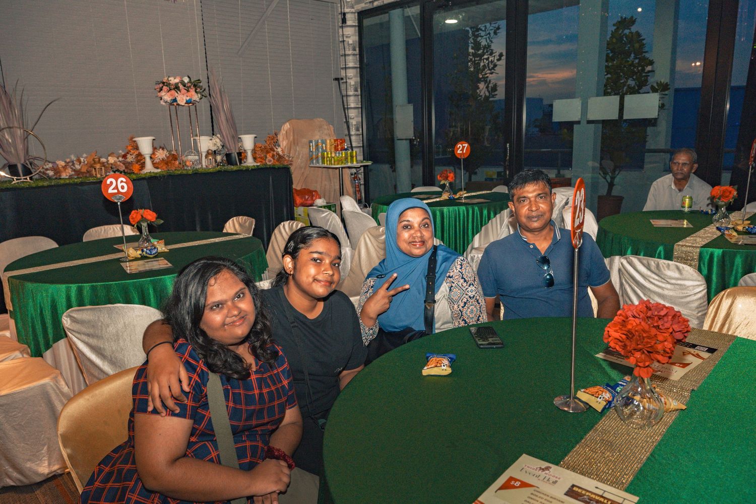 Guests seated at tables with green tablecloths and autumn-themed centerpieces at an indoor event.