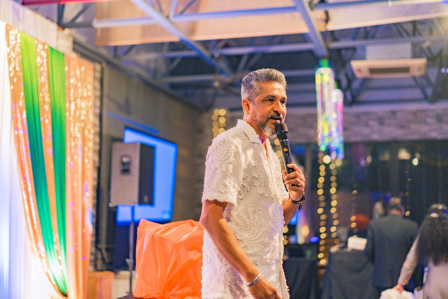 Speaker in white shirt addresses audience at decorated venue with colored lighting.