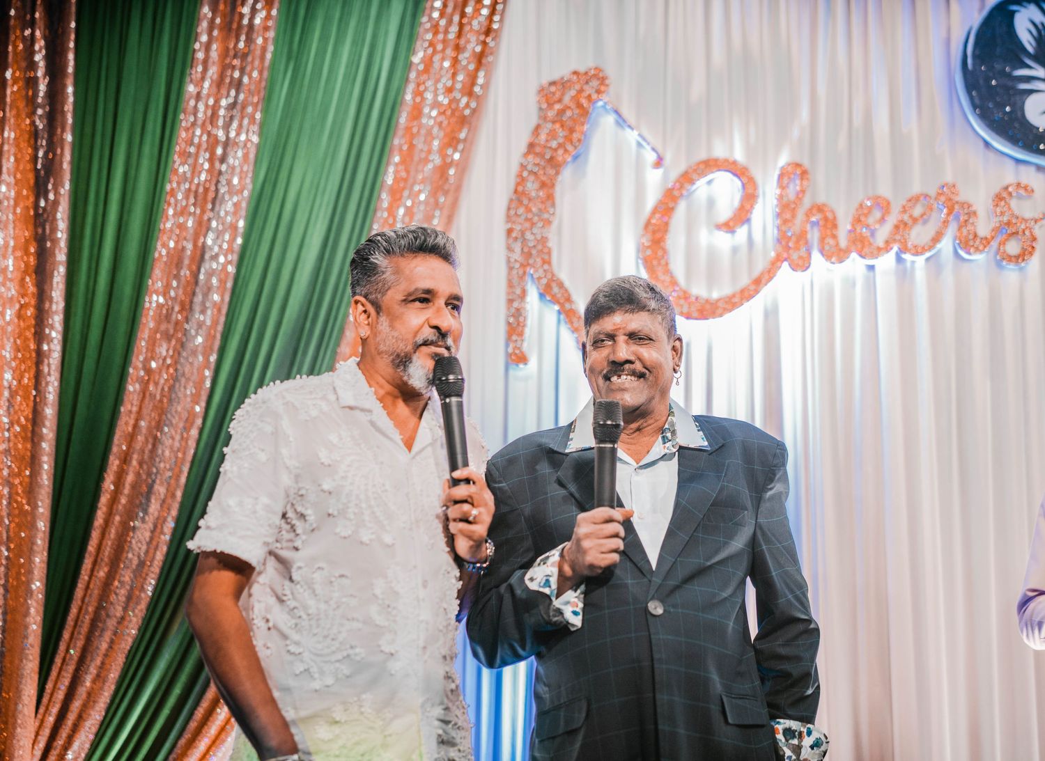 Two men with microphones performing on stage with sparkling backdrop.