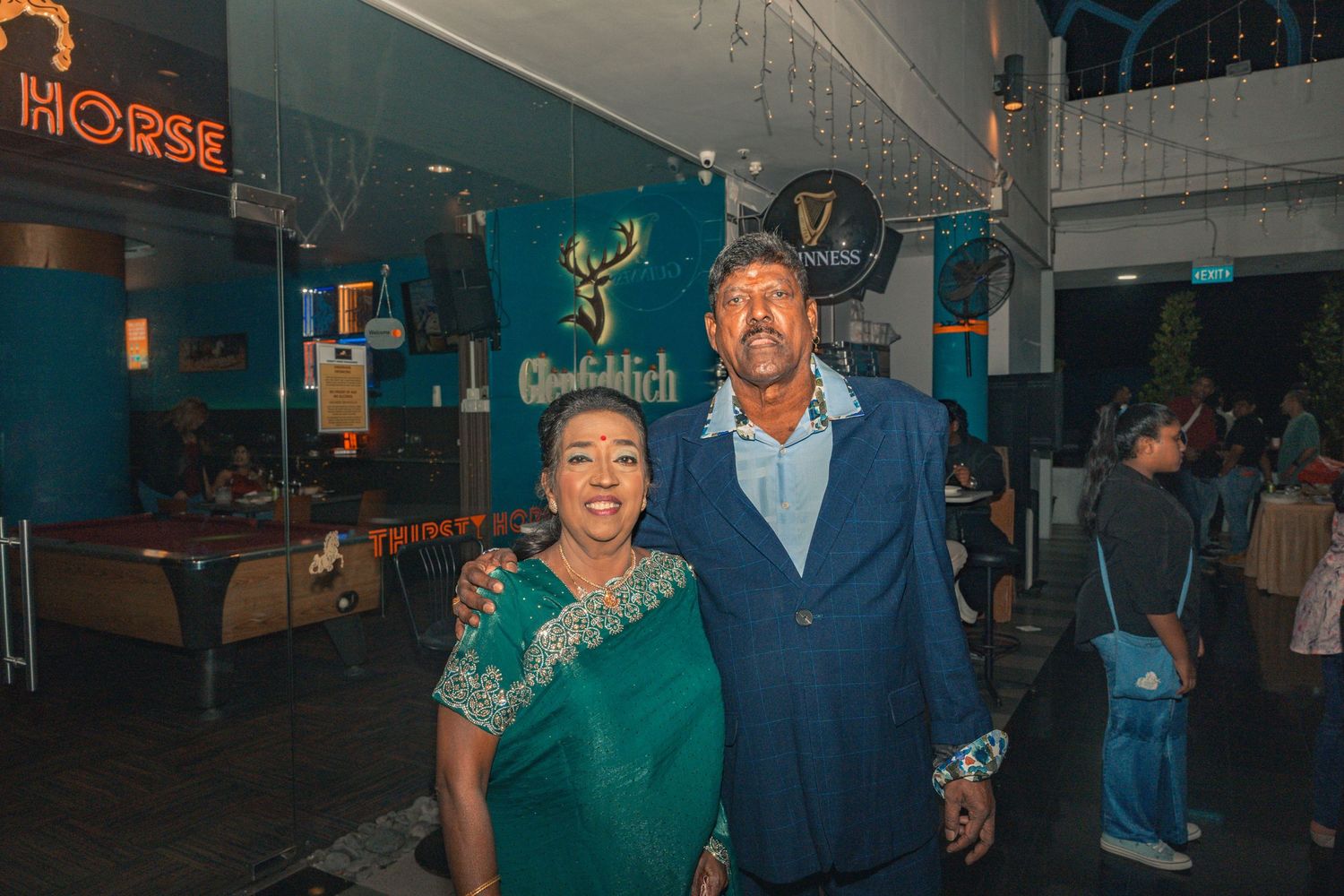 Two people posing together at a venue with blue walls and neon signage.