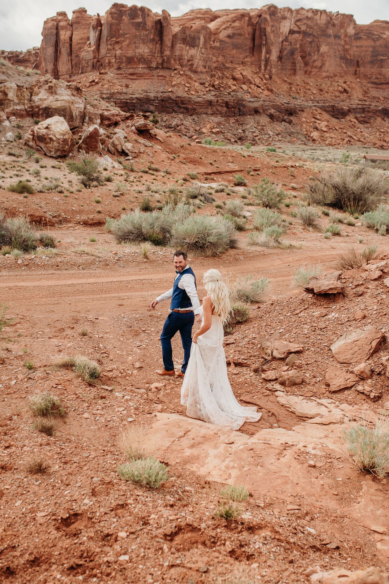 Wedding couple walking along desert trail with dramatic red rock cliffs in the background.