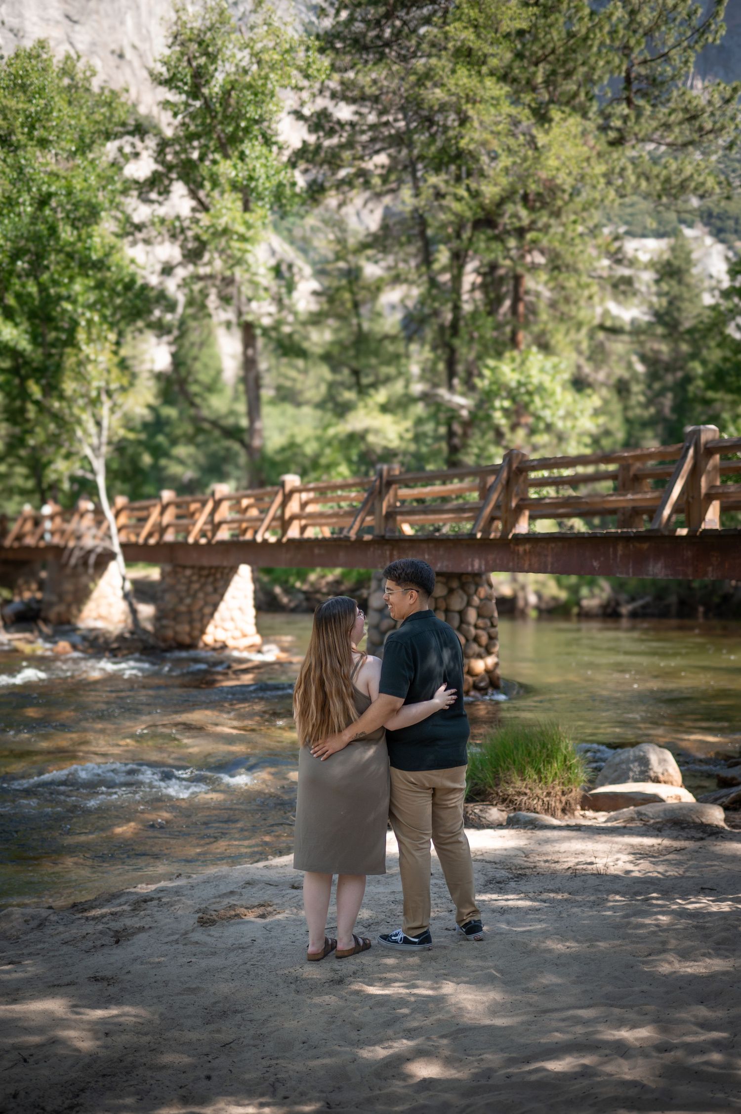 A newly engaged couple embrace on a beach by a bridge in Yosemite