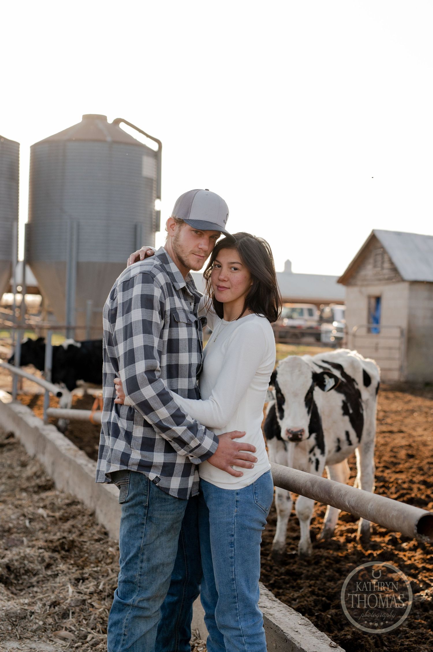 A couple shares an intimate moment on their farm with dairy cows and grain silos behind them.