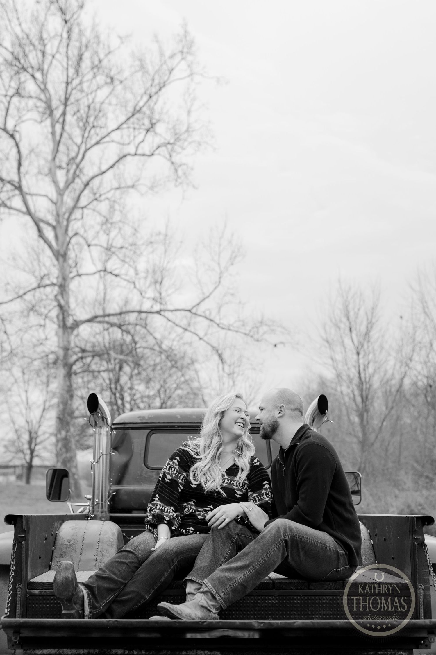 A romantic black and white photo of a couple sitting in the bed of a vintage pickup truck on a winter day.