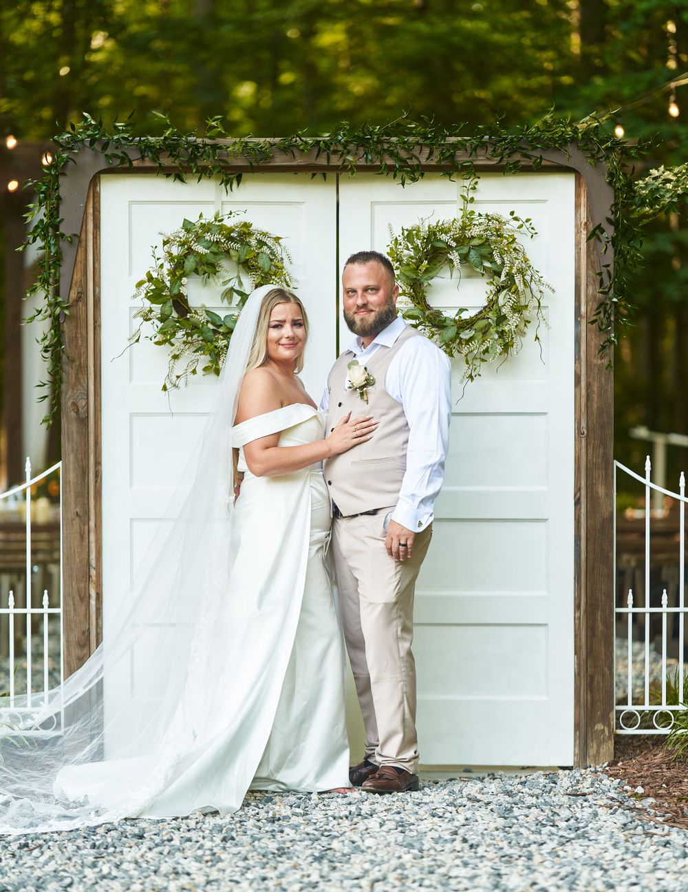 Newlywed couple poses against white doors decorated with green wreaths in outdoor setting.