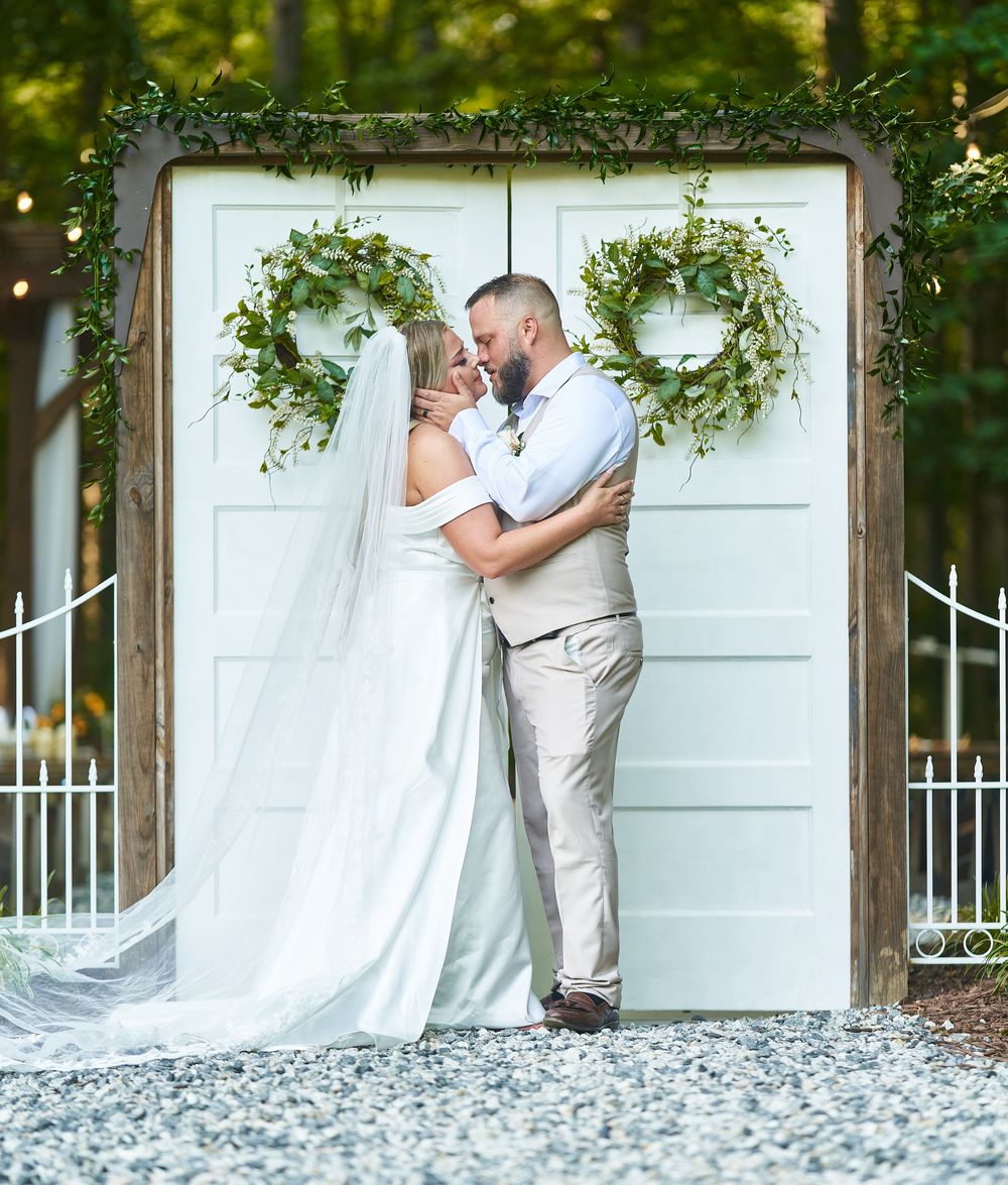 Bride and groom sharing a kiss in front of rustic white doors adorned with greenery wreaths at an outdoor wedding.