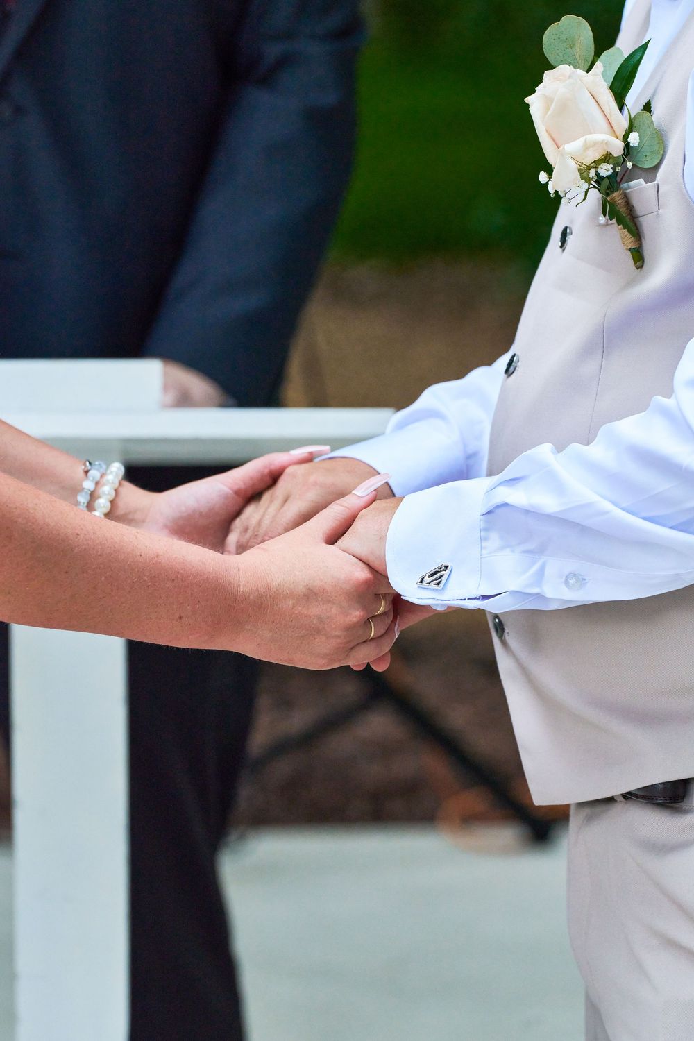 Bride and groom holding hands during outdoor wedding ceremony, groom wearing white suit with rose boutonniere.