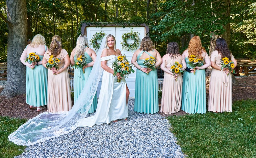 Bridesmaids in mint and blush dresses pose with bride holding sunflower bouquets on gravel path.