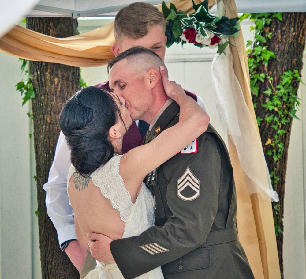 Military groom in uniform shares first kiss with bride at outdoor wedding ceremony under floral arch.