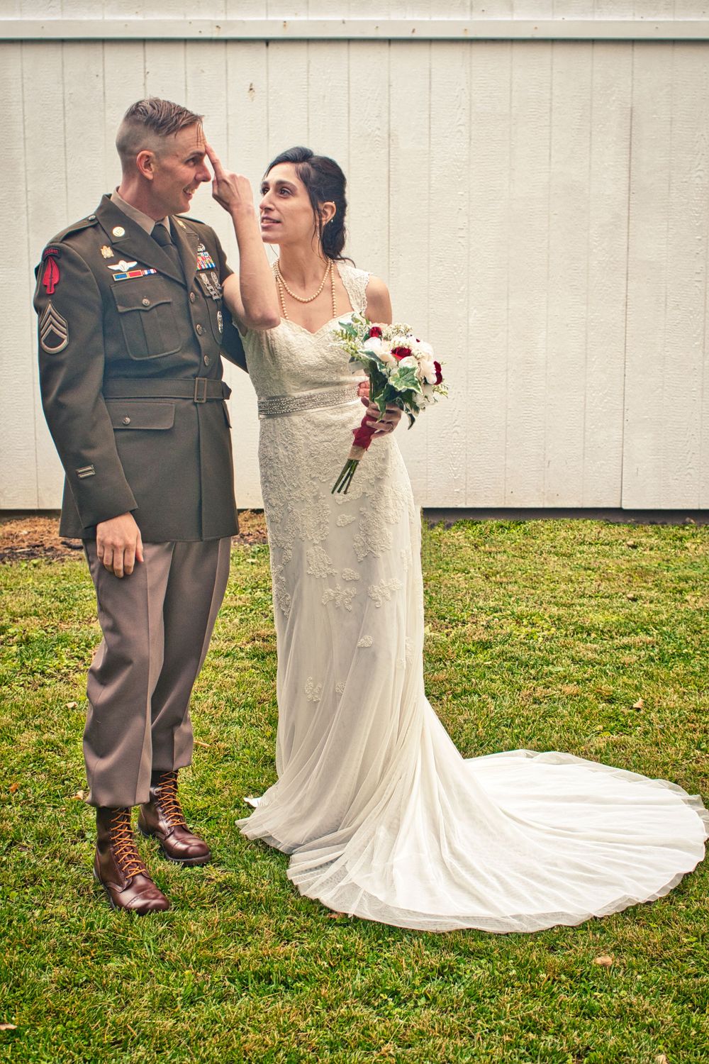 Military groom in uniform and bride in white gown share tender moment outdoors, bride touching grooms face while holding bouquet.