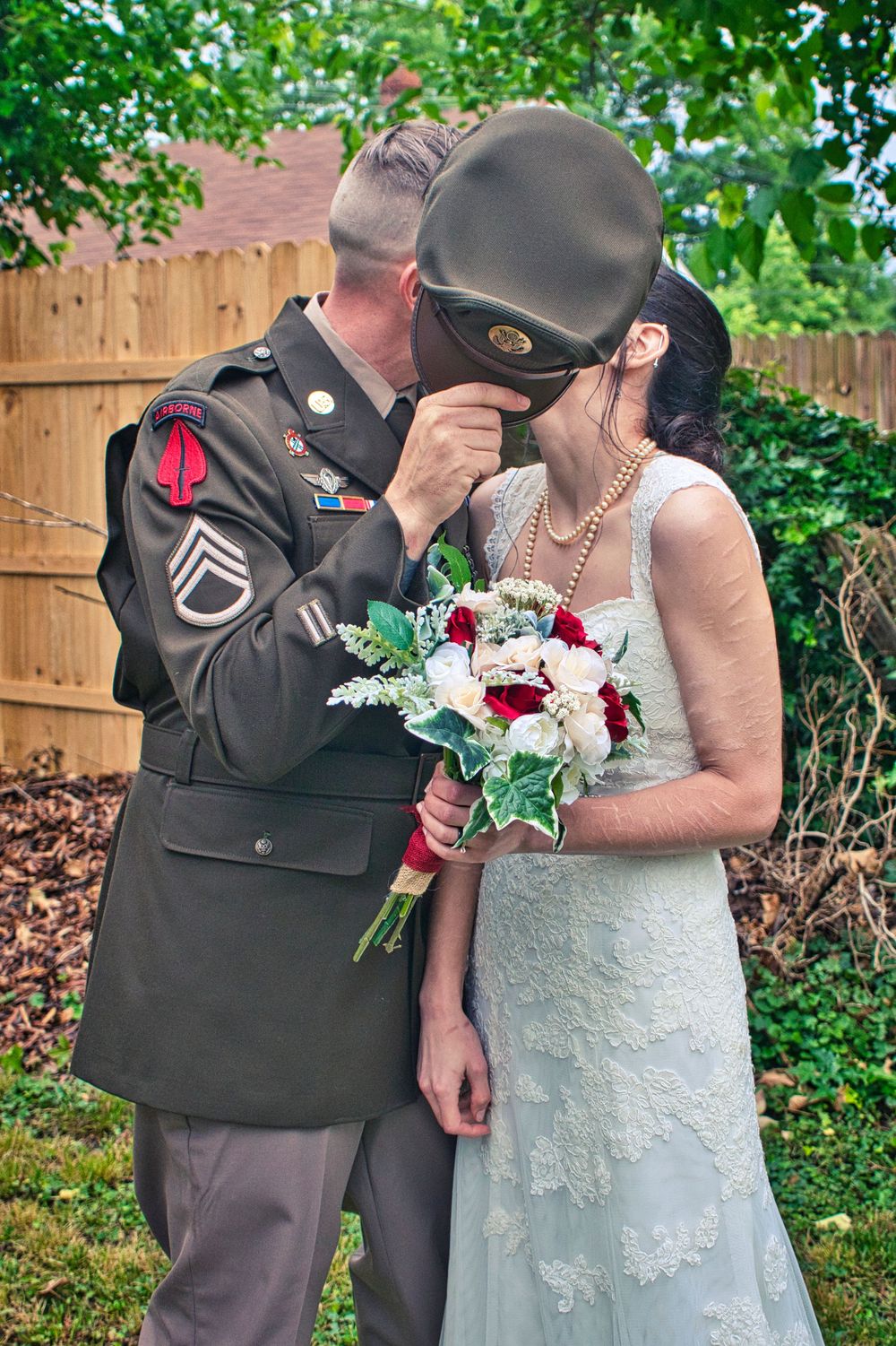Military groom in dress uniform kissing bride holding red and white bouquet at outdoor wedding ceremony.