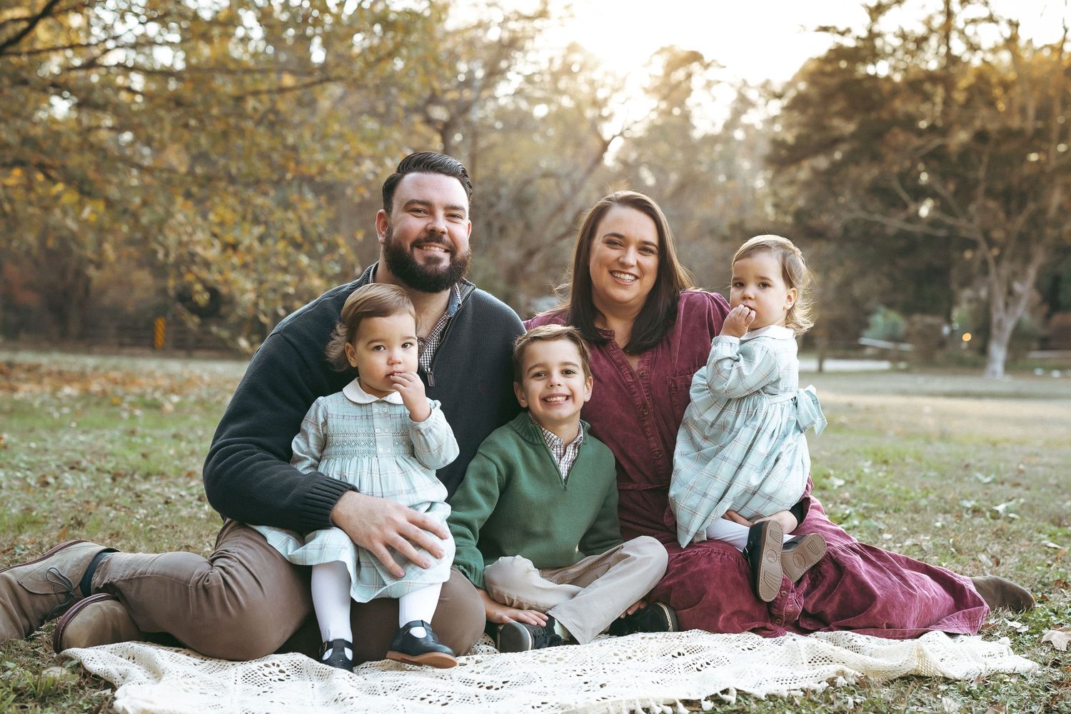 A family sits together on a blanket outdoors during golden hour in an autumn setting.