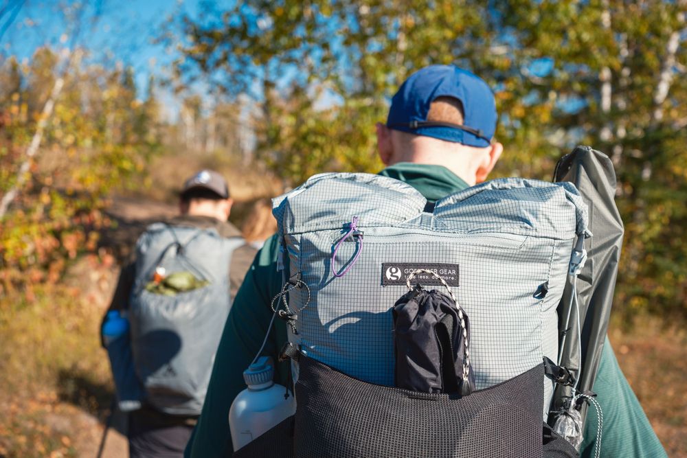 Hikers wearing backpacks trek along a scenic mountain trail surrounded by autumn foliage and blue skies.