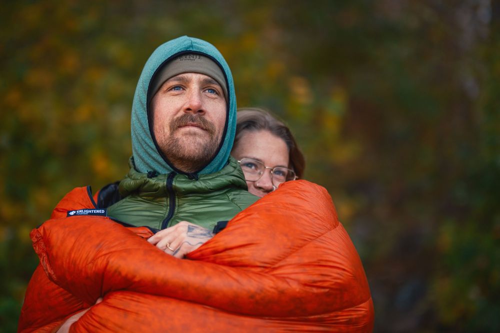 Two people snuggle in bright orange sleeping bags while camping outdoors during fall weather.