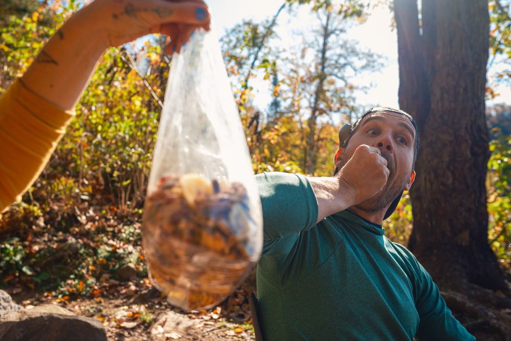 A person pours water from a clear plastic bottle into a small cup while standing in a wooded outdoor setting.