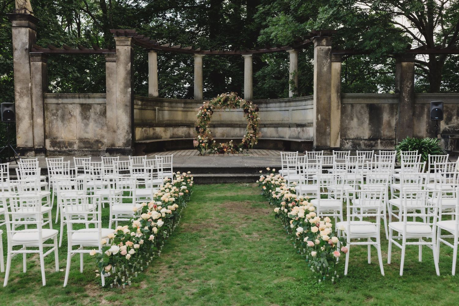 Rows of white garden chairs arranged outdoors for a wedding ceremony with stone pillars and greenery in the background.