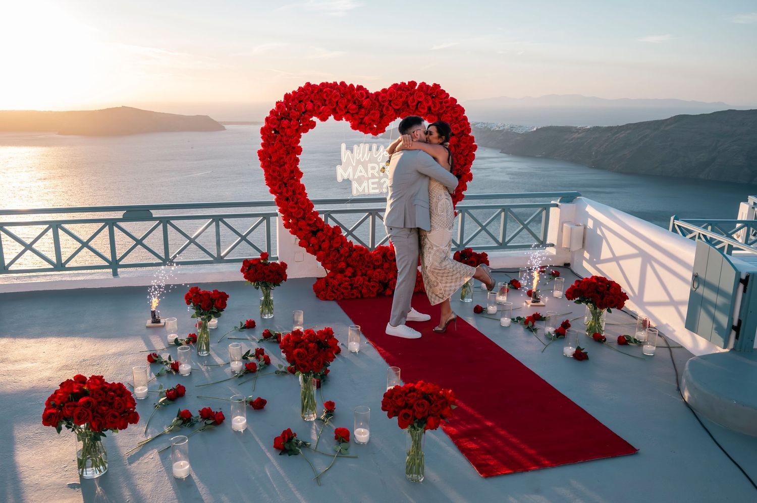 Elegant proposal setup with red rose heart arch and red carpet overlooking Santorini caldera.