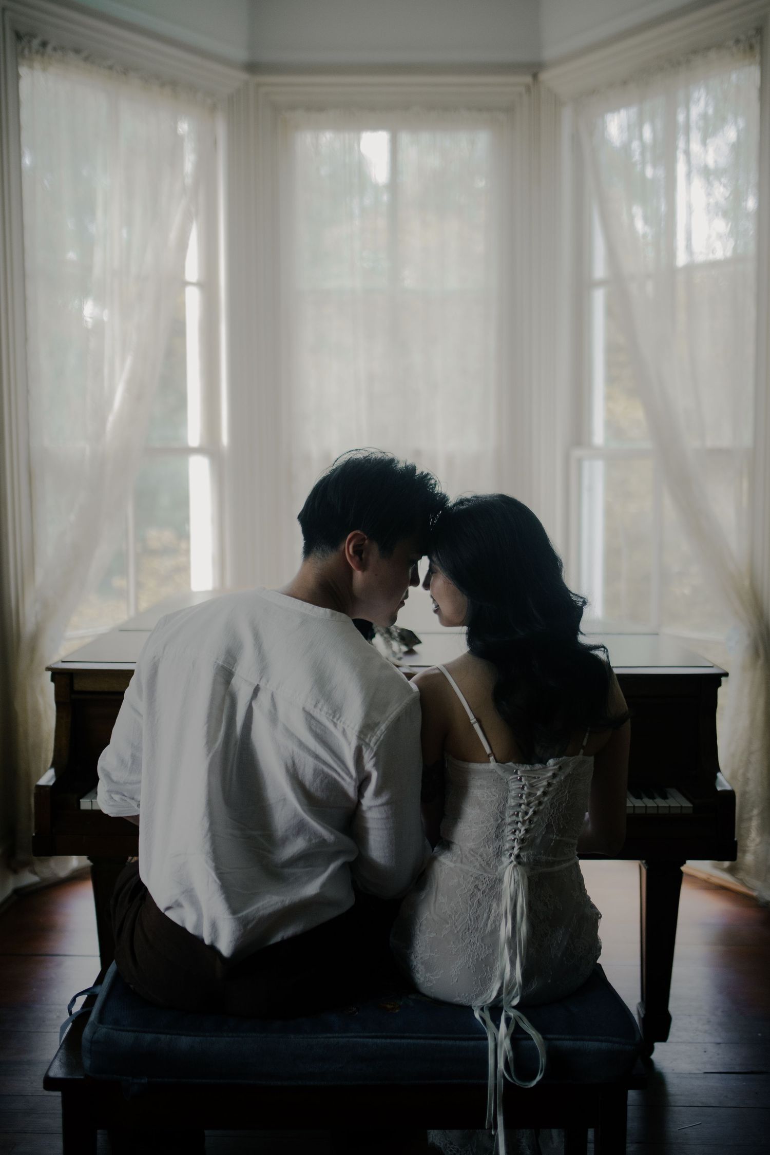 A couple sits close together on a bench in front of sheer curtained windows.