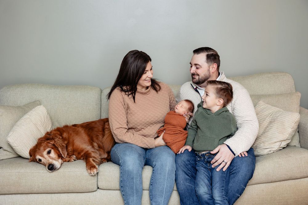 Family with newborn, toddler, and golden retriever sitting together on a cozy beige sofa.