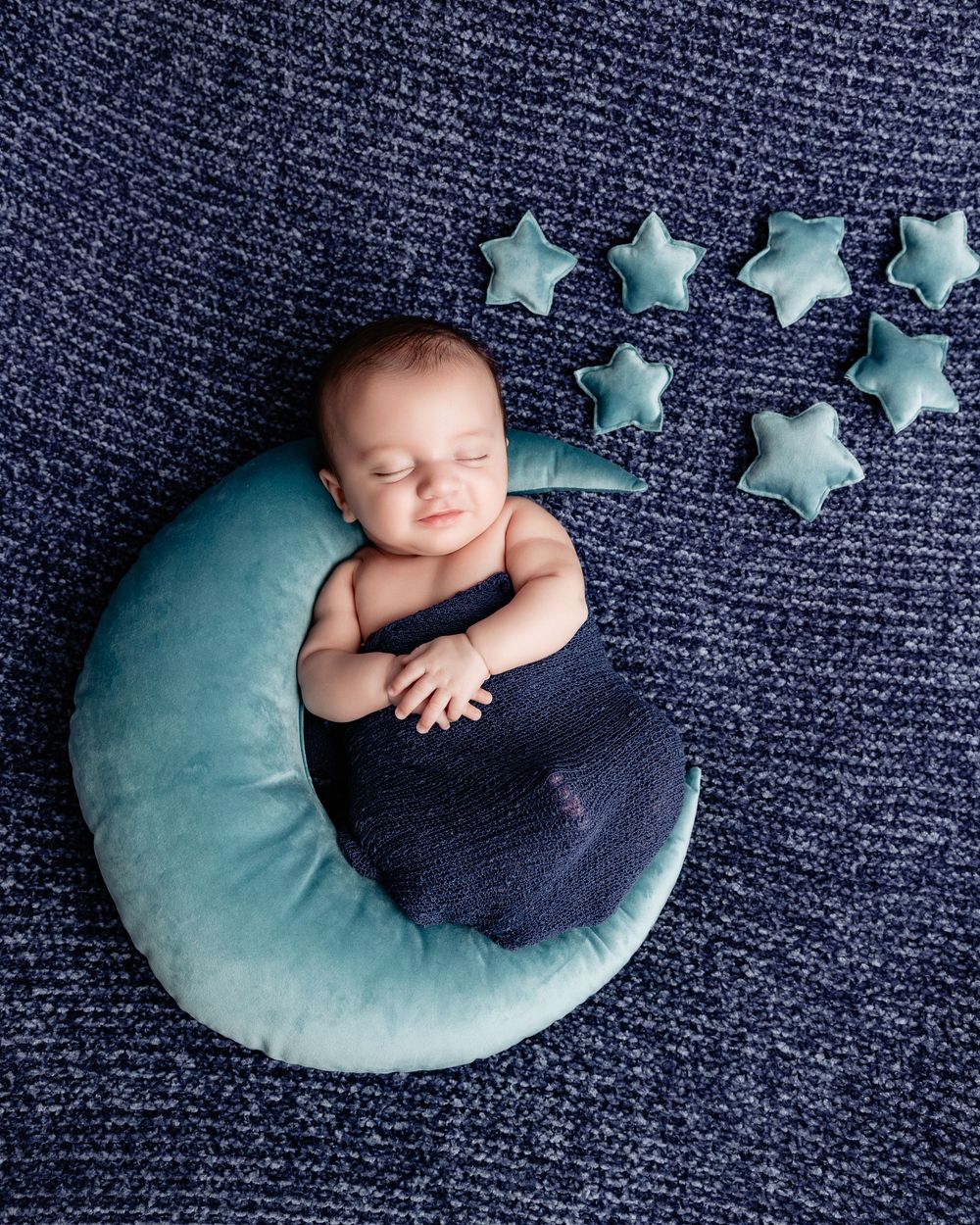 Newborn baby sleeping on a teal crescent moon pillow surrounded by star cushions on a navy blue background.