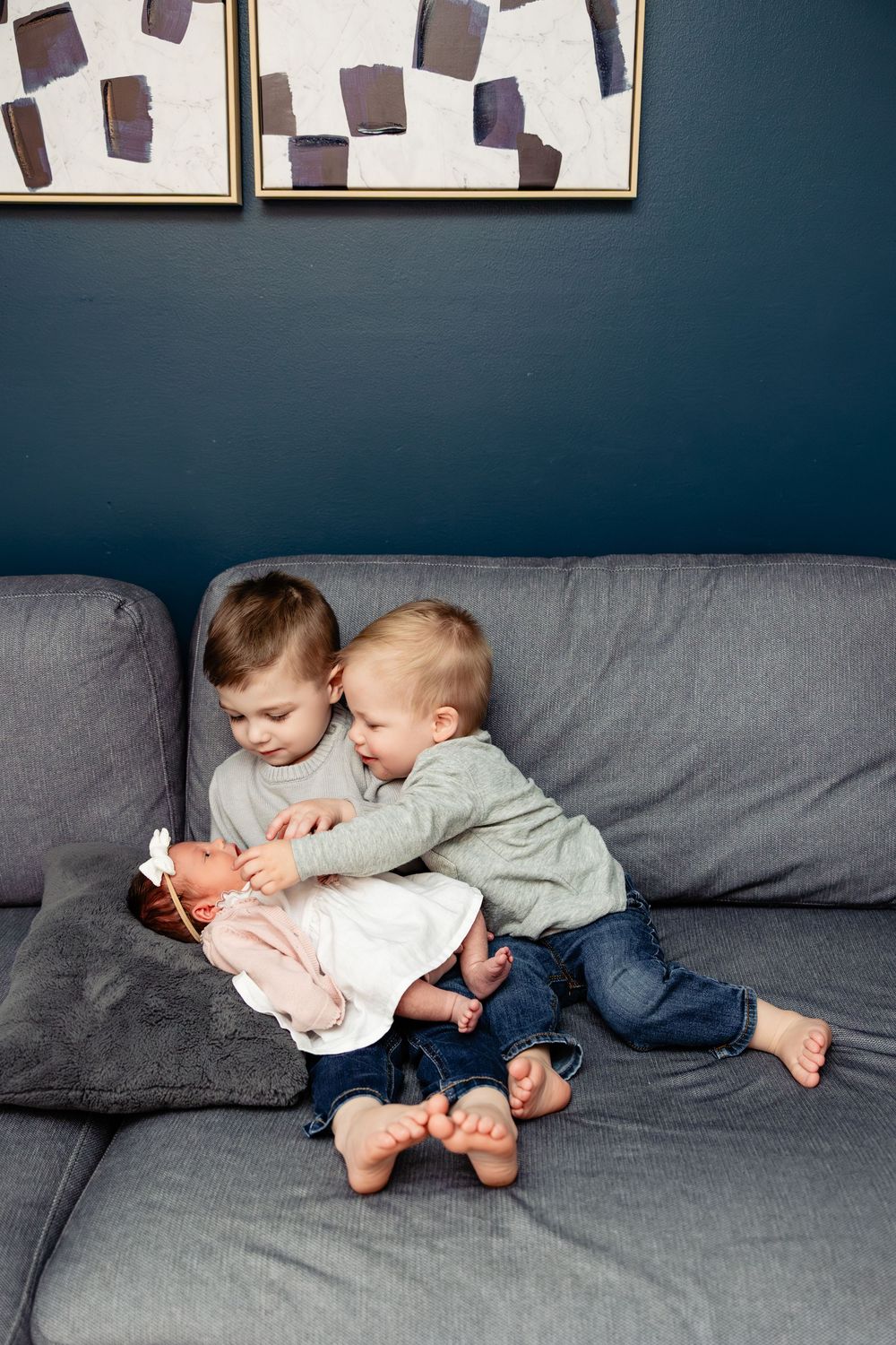 Two young siblings sitting on a gray couch, gently holding a newborn baby in a cozy home setting.