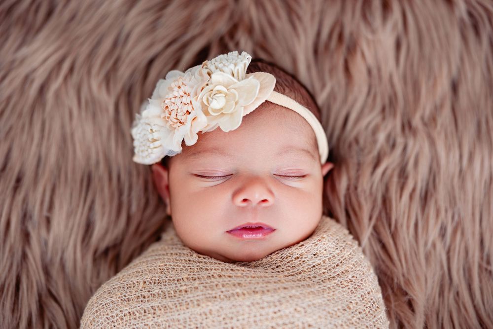 Sleeping newborn girl swaddled in burlap wrap wearing a lace floral headband on fur backdrop.