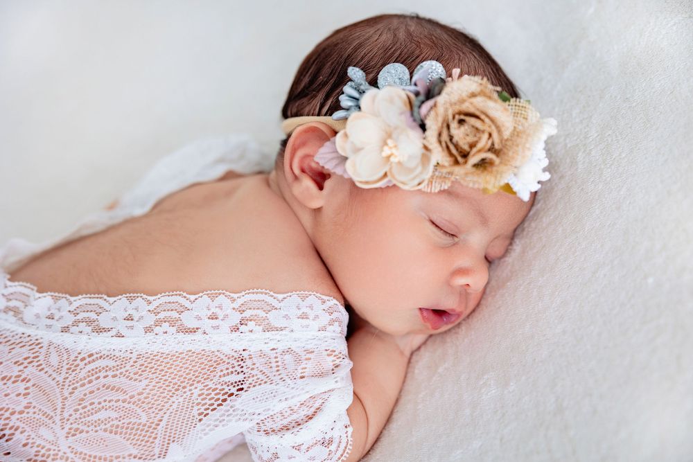 Peaceful newborn baby girl sleeping on white fur wearing a floral headband and lace wrap.