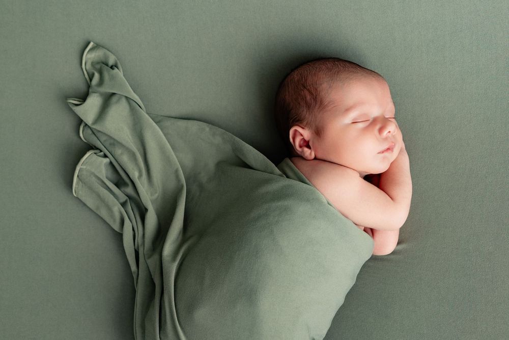 Newborn baby swaddled in sage green wrap sleeping peacefully on matching green backdrop.