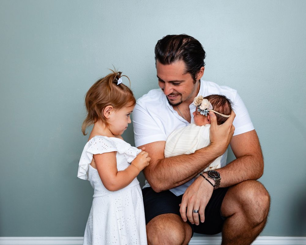 Father crouching and holding newborn while young daughter in white dress looks on adoringly.