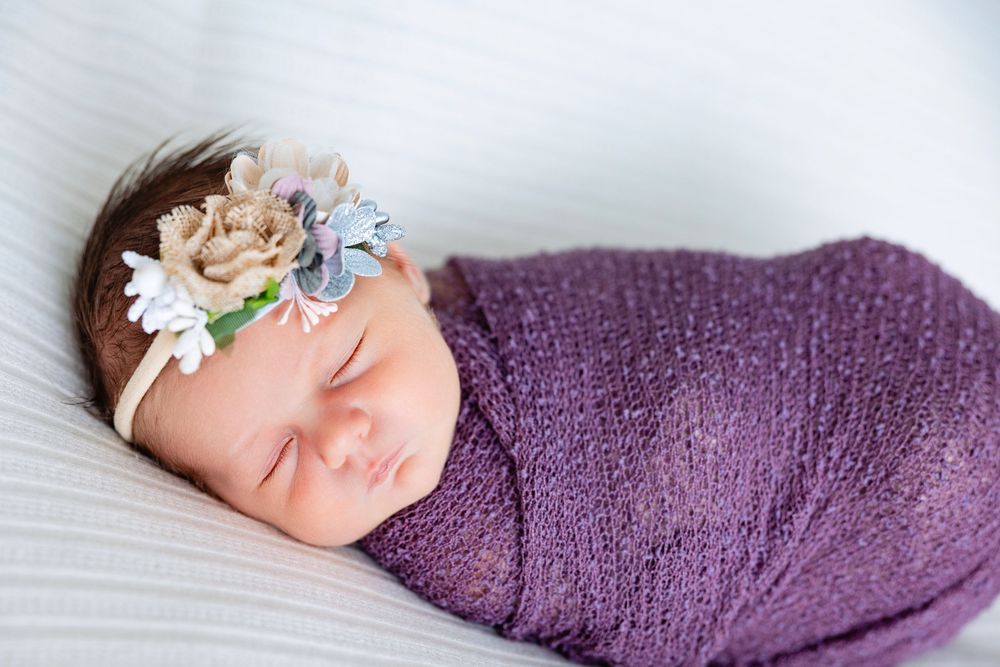 Sleeping newborn baby girl swaddled in purple wrap wearing a floral headband on white backdrop.