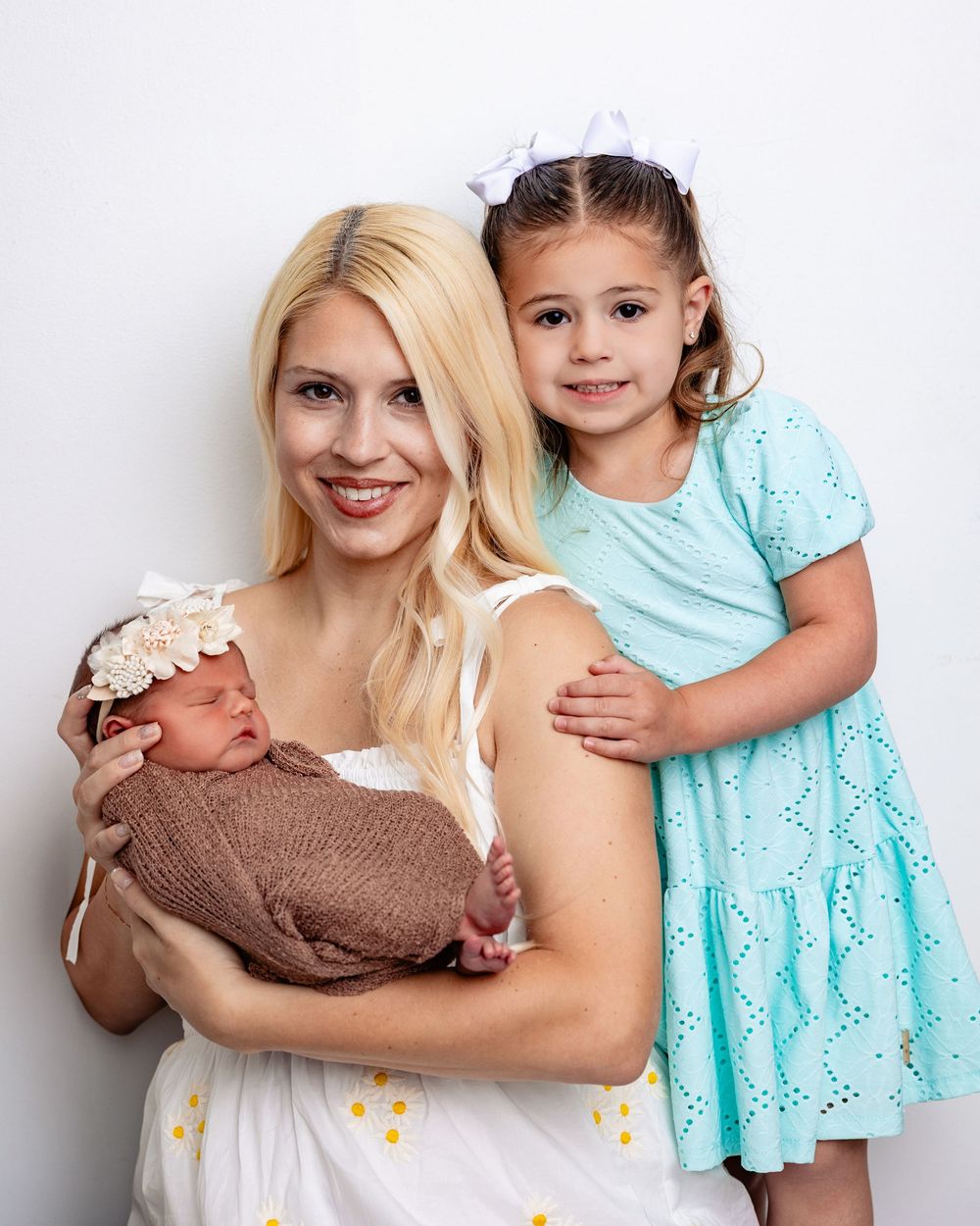 Smiling blonde mother holding newborn baby while older daughter in teal dress poses beside her.