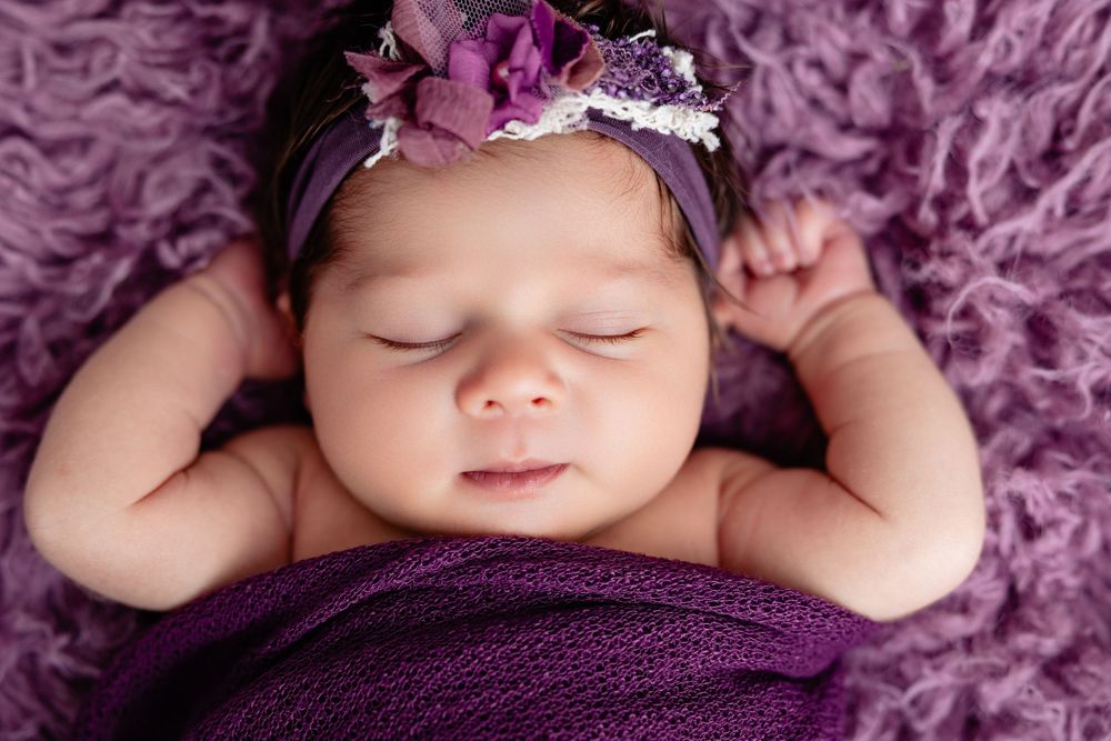 Peaceful newborn baby girl sleeping wrapped in purple, wearing a floral headband on a soft lavender blanket.