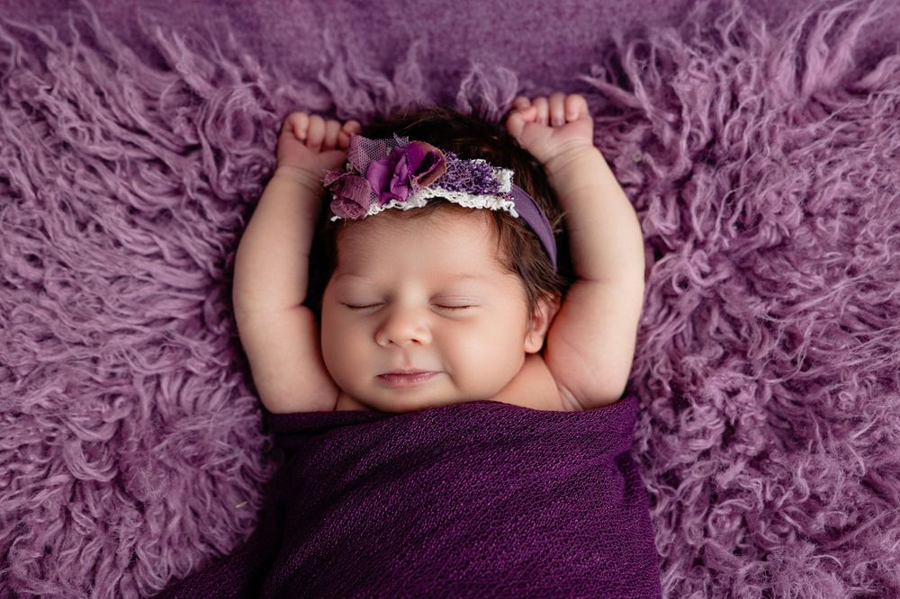 Sleeping newborn baby girl swaddled in purple wrap with floral headband on fluffy rug.