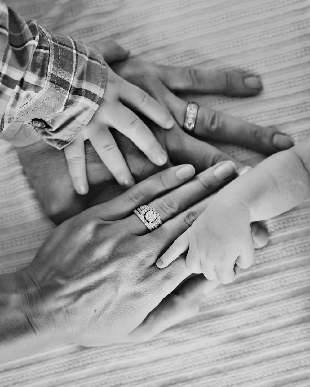 Family hands stacked together showing wedding rings and a tiny newborn baby hand on a wooden surface.