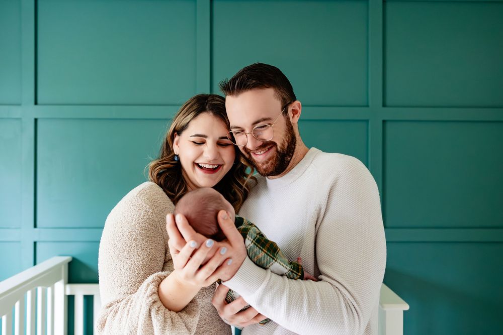 Smiling parents joyfully looking at their newborn baby in front of a teal paneled wall.